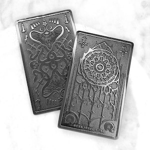 Può includere: Due targhe rettangolari in metallo grigio scuro con disegni in rilievo intricati. Una presenta un motivo di serpente e spada, l'altra un disegno di vetrata con un occhio. La frase "MORS IANUA VITAE" è in basso su una targa.