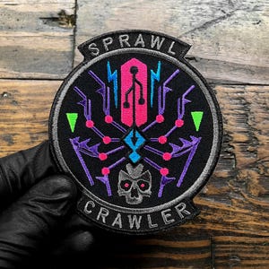 Puede incluir: Un parche redondo negro con las palabras "SPRAWL CRAWLER" en blanco. El parche presenta un dise&ntilde;o de ara&ntilde;a estilizado en rosa, morado, azul y verde, con una calavera en la parte inferior. El parche est&aacute; sujeto por una mano enguantada.