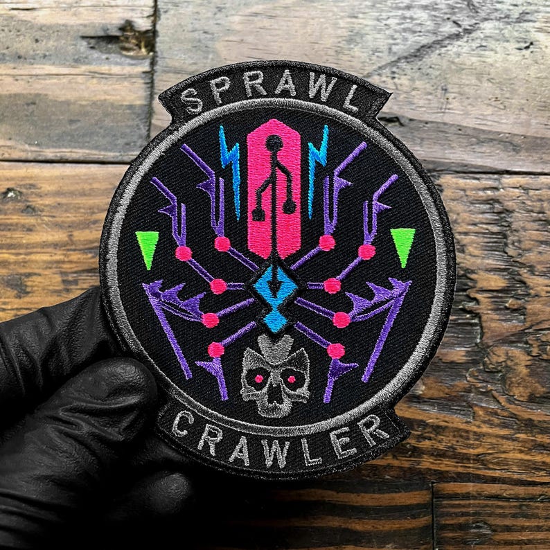 Puede incluir: Un parche redondo negro con las palabras "SPRAWL CRAWLER" en blanco. El parche presenta un dise&ntilde;o de ara&ntilde;a estilizado en rosa, morado, azul y verde, con una calavera en la parte inferior. El parche est&aacute; sujeto por una mano enguantada.