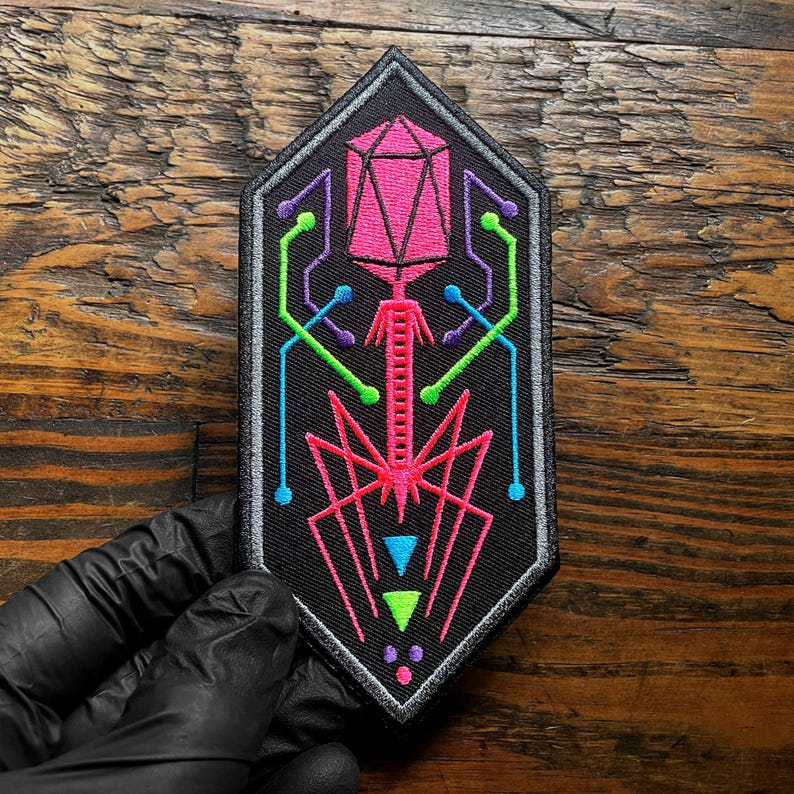 May include: Black embroidered patch featuring a geometric design in neon pink, green, blue, and purple. The patch has a grey border and an unusual shape. The design appears to be a stylized representation of a virus or similar scientific concept.