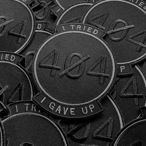 K&ouml;nnte beinhalten: Eine Ansammlung schwarzer, gestickter Aufn&auml;her. Jeder runde Aufn&auml;her zeigt die durchgestrichene Zahl 404 und die wei&szlig;en Schriftz&uuml;ge &bdquo;I Tried&ldquo; und &bdquo;I Gave Up&ldquo;. Die Aufn&auml;her sind mit einem wei&szlig;en Rand versehen.