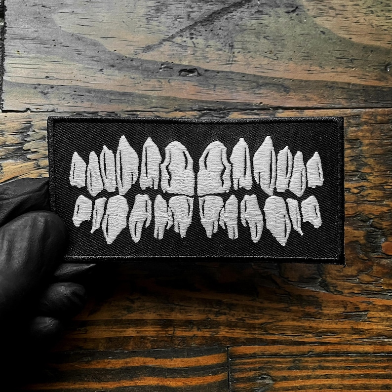 K&ouml;nnte beinhalten: Ein schwarzer rechteckiger Aufn&auml;her mit einem wei&szlig;en, gestickten Design von Z&auml;hnen. Die Z&auml;hne sind in zwei Reihen angeordnet, die den Ober- und Unterkiefer darstellen. Der Aufn&auml;her ist zum Aufn&auml;hen auf Kleidung oder Accessoires gedacht.
