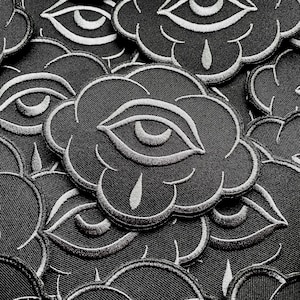 K&ouml;nnte beinhalten: Nahaufnahme von schwarzen, wolkenf&ouml;rmigen, gestickten Aufn&auml;hern mit einem wei&szlig;en Augendesign. Die Aufn&auml;her sind dicht angeordnet und &uuml;berlappen sich. Jedes Augendesign enth&auml;lt ein Tr&auml;nendetail.