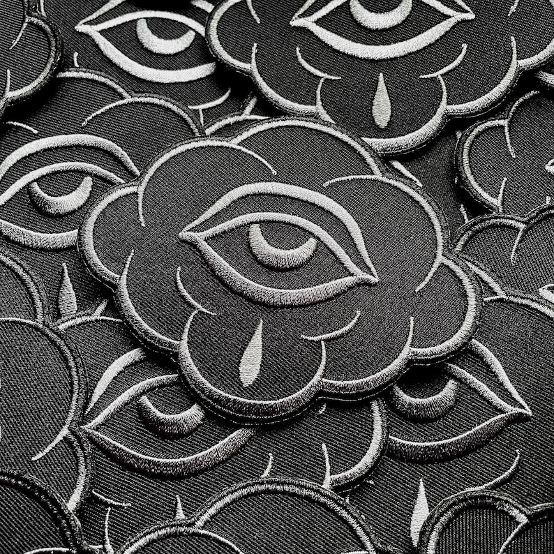 K&ouml;nnte beinhalten: Nahaufnahme von schwarzen, wolkenf&ouml;rmigen, gestickten Aufn&auml;hern mit einem wei&szlig;en Augendesign. Die Aufn&auml;her sind dicht angeordnet und &uuml;berlappen sich. Jedes Augendesign enth&auml;lt ein Tr&auml;nendetail.