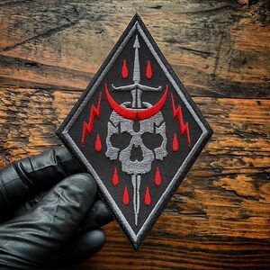 May include: A diamond-shaped embroidered patch featuring a grey skull pierced by a vertical sword. A red crescent moon sits above the skull, flanked by two red lightning bolts. Small red teardrop shapes are scattered around the central design.
