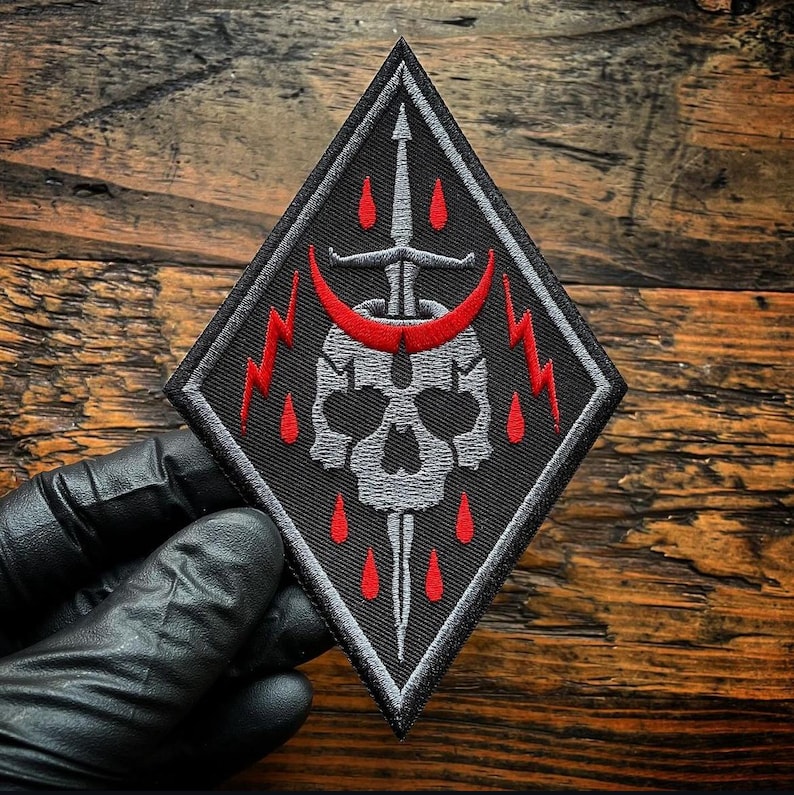May include: A diamond-shaped embroidered patch featuring a grey skull pierced by a vertical sword. A red crescent moon sits above the skull, flanked by two red lightning bolts. Small red teardrop shapes are scattered around the central design.