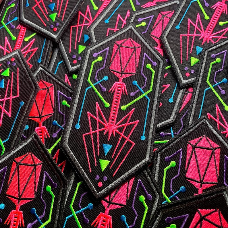 May include: A pile of black embroidered patches with a geometric design. Each patch features a pink, geometric shape with a stem, surrounded by neon green, blue, and purple lines. The patches have a grey border.