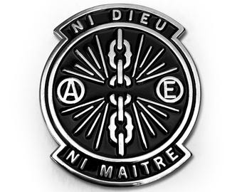 May include: A black and silver metal pin with the words "NI DIEU NI MAITRE" and a central design featuring chains and radiating lines. The pin also includes the letters "A" and "E".