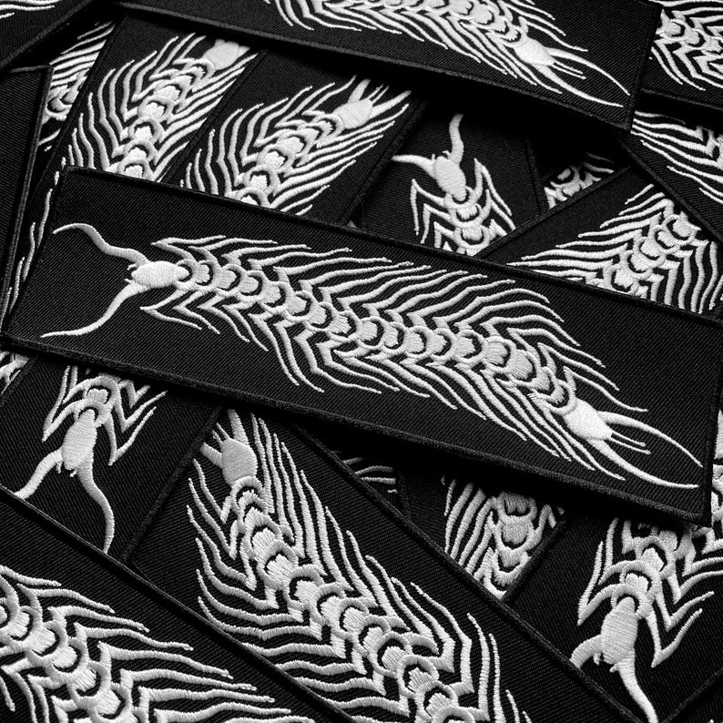 May include: A collection of black rectangular patches featuring a white embroidered dragon motif. The dragon has a detailed, flowing design. The patches are arranged in a layered pattern. These patches could be used to decorate clothing or accessories.