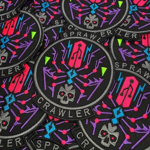 Puede incluir: Parches redondos negros bordados con un dise&ntilde;o de calavera y telara&ntilde;a. Los parches presentan la palabra "CRAWLER" y un s&iacute;mbolo USB estilizado en rosa y azul. Acentos en morado, verde y gris.