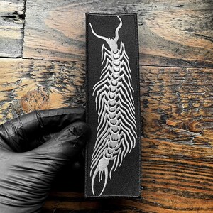 May include: Black rectangular patch with a white centipede design. The embroidered insect has a segmented body and curved antennae. The patch is designed for clothing or accessories.