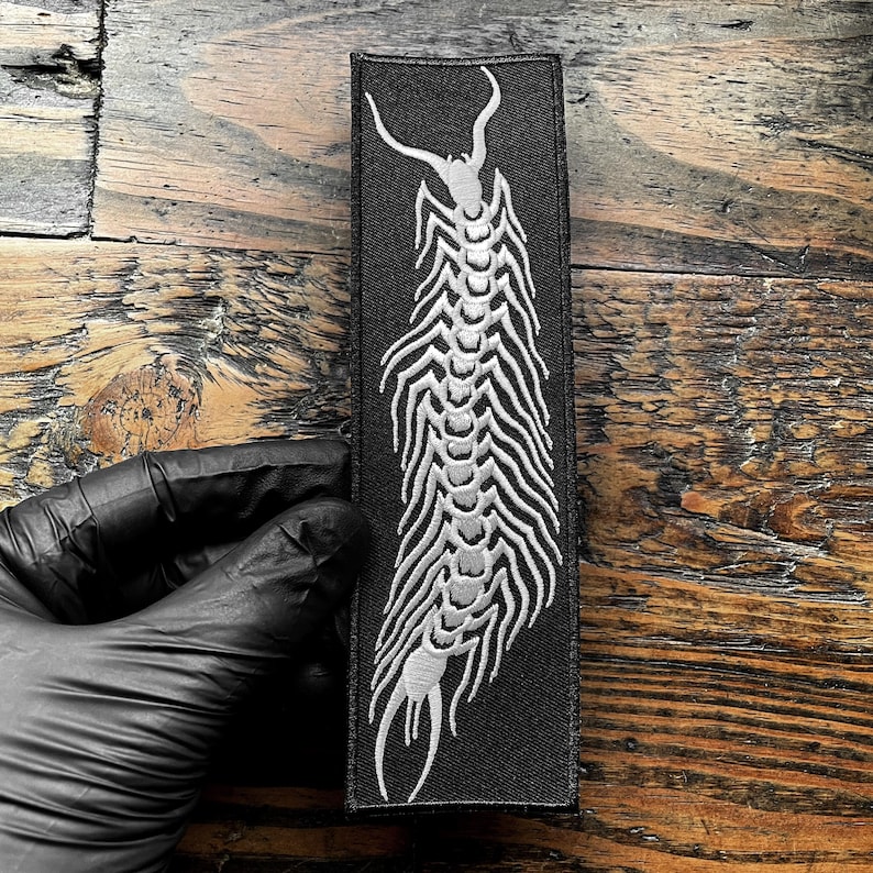 May include: Black rectangular patch with a white centipede design. The embroidered insect has a segmented body and curved antennae. The patch is designed for clothing or accessories.