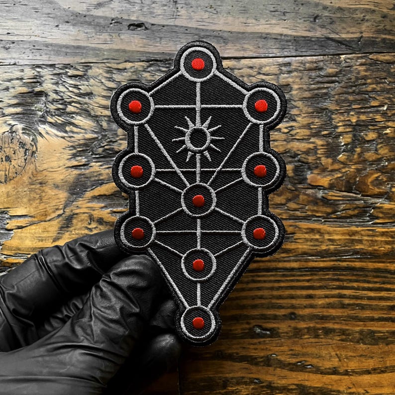 May include: Black embroidered patch featuring a geometric design with red circles and a sunburst in the center. The patch has a white border and is likely for clothing or accessories.