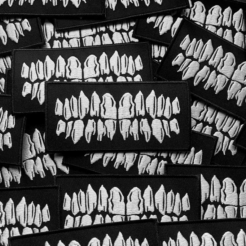 K&ouml;nnte beinhalten: Eine Ansammlung schwarzer rechteckiger Aufn&auml;her mit einem wei&szlig;en, gestickten Zahnmotiv. Die Aufn&auml;her sind verstreut, einige &uuml;berlappen sich. Das Zahnmotiv ist detailliert und zeigt die oberen und unteren Zahnreihen.