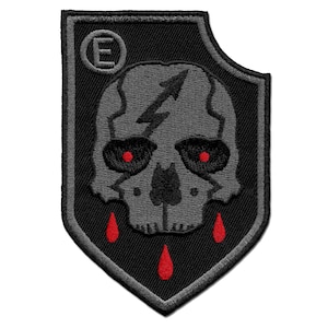 Può includere: Toppa ricamata nera e grigia con un designo di teschio e ossa incrociate. Il teschio ha occhi rossi e gocciola sangue. La toppa ha la forma di uno scudo nero con una lettera "E" bianca in un cerchio nell'angolo in alto a sinistra.