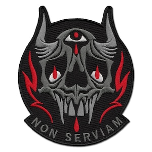 Puede incluir: Parche bordado con un diseño de calavera estilizada, cuernos y un ojo, con detalles rojos y blancos sobre fondo negro. El texto "NON SERVIAM" está en la parte inferior. El parche es de forma circular.