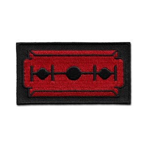 May include: A rectangular black patch with a red razor blade design. The razor blade is outlined in black and features a series of black circles and diamond shapes. The patch is likely an iron-on or sew-on accessory.