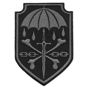 May include: A black and grey embroidered patch in a shield shape. The design includes an umbrella, raindrops, a sword, crossed bones, and chains. The patch has a detailed, textured appearance.