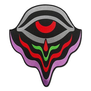 May include: A black patch with a stylized eye design. The eye features a red iris, gray and black striped eyelid, and a series of red, green, and purple wavy lines below. The patch has a unique, flowing shape.
