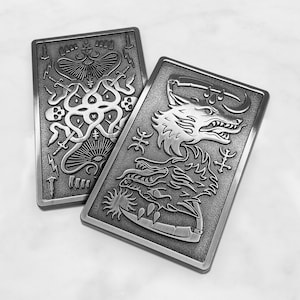 Puede incluir: Dos placas rectangulares de metal plateado con intrincados diseños en relieve. Una presenta una criatura parecida a un pulpo con espadas y rayos. La otra representa un lobo y un dragón con símbolos estilizados. Las placas son probablemente decorativas o funcionales.