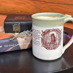 Può includere: Una tazza in ceramica color crema con bordo verde chiaro. Presenta un'illustrazione color amaranto di un angelo e del testo. Un libro, una croce e un altro libro sono accanto alla tazza.