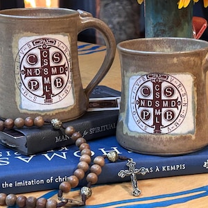 May include: Two ceramic mugs with handles, featuring a religious emblem in a white square. The mugs are brown with a textured finish. A rosary and a crucifix rest on a stack of books, including "The Imitation of Christ."