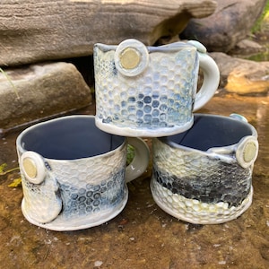 May include: Three ceramic mugs with a textured, honeycomb-like design. The mugs are white with blue and black accents and have gold accents. The mugs are stacked on top of each other.