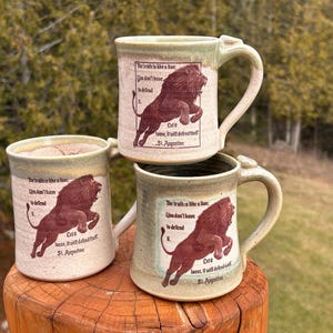 May include: Three ceramic mugs with a speckled beige finish and a green rim. Each mug features a dark red illustration of a lion and the text: The truth is like a lion; You don't have to defend it. Let it loose, it will defend itself. St. Augustine.