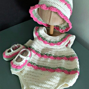 Puede incluir: Un conjunto de ropa de bebé tejido a crochet en blanco, rosa y verde. El conjunto incluye un gorro, un mono y un par de botines.