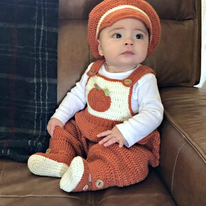 May include: A baby wearing a crocheted orange and white pumpkin-themed outfit with a matching crocheted hat. The baby is sitting on a brown leather chair.