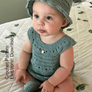 May include: A baby wearing a crocheted outfit in a light blue color. The outfit includes a hat, a sleeveless dress, and shoes. The dress and shoes have pink flower accents.