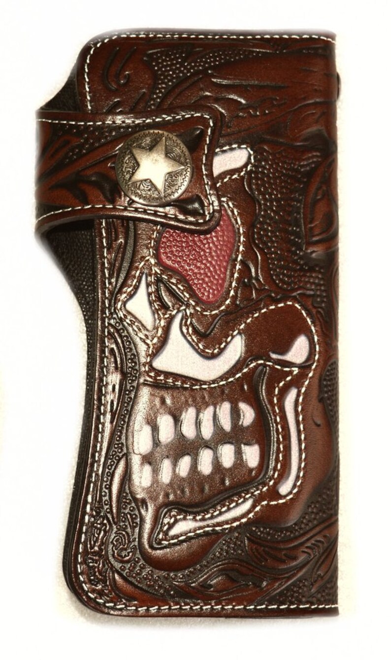 Puede incluir: Una cartera de cuero marr&oacute;n con un dise&ntilde;o de calavera. La calavera es blanca con la nariz roja y est&aacute; rodeada de intrincados patrones florales. La cartera tiene un bot&oacute;n de metal en forma de estrella.