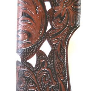 Puede incluir: Una cartera de cuero marr&oacute;n con un dise&ntilde;o floral tallado. La cartera tiene costuras blancas alrededor de los bordes y un cierre de presi&oacute;n plateado.