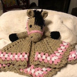 May include: A crochet lovey featuring a tan horse head with a black mane, black hooves, and a pink halter. The attached blanket is made of tan yarn with a decorative border of pink and white crochet clusters, creating a soft, textured appearance.
