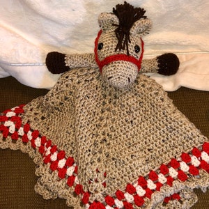 May include: A crocheted horse security blanket featuring a tan horse head with a dark brown mane, black eyes, and a red bridle. The attached blanket is tan with a decorative border of red and white crocheted clusters along the outer edges.