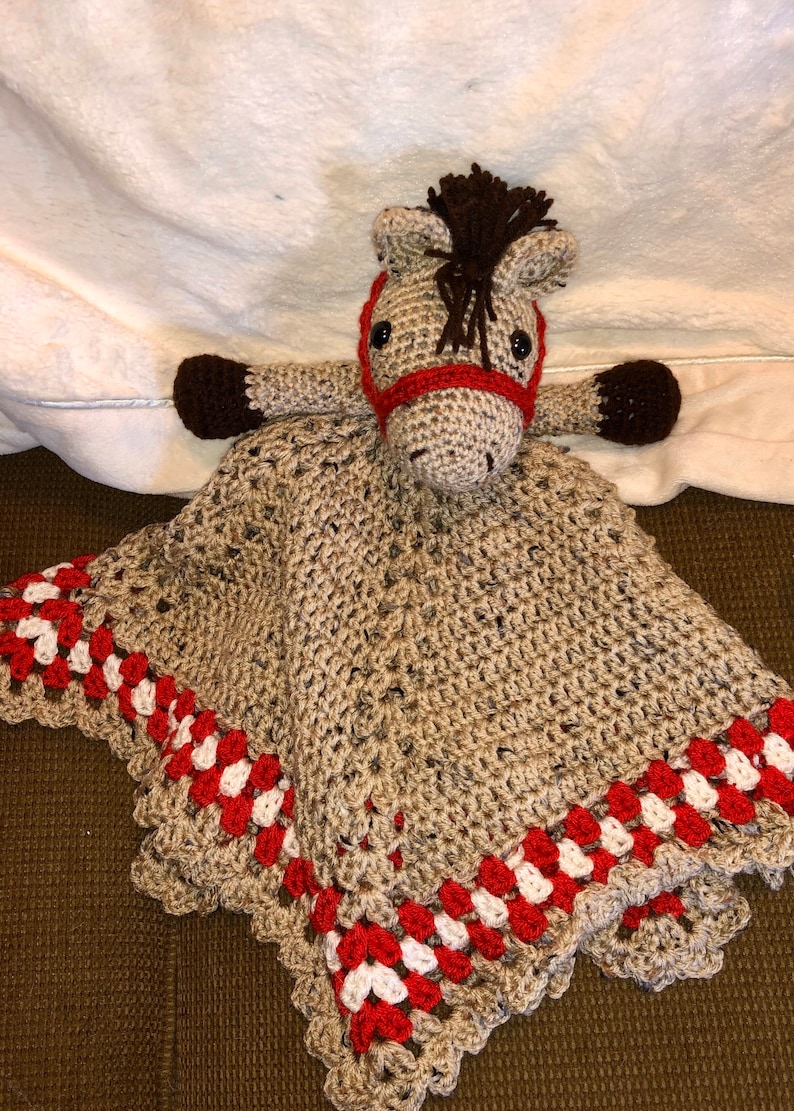 May include: A crocheted horse security blanket featuring a tan horse head with a dark brown mane, black eyes, and a red bridle. The attached blanket is tan with a decorative border of red and white crocheted clusters along the outer edges.