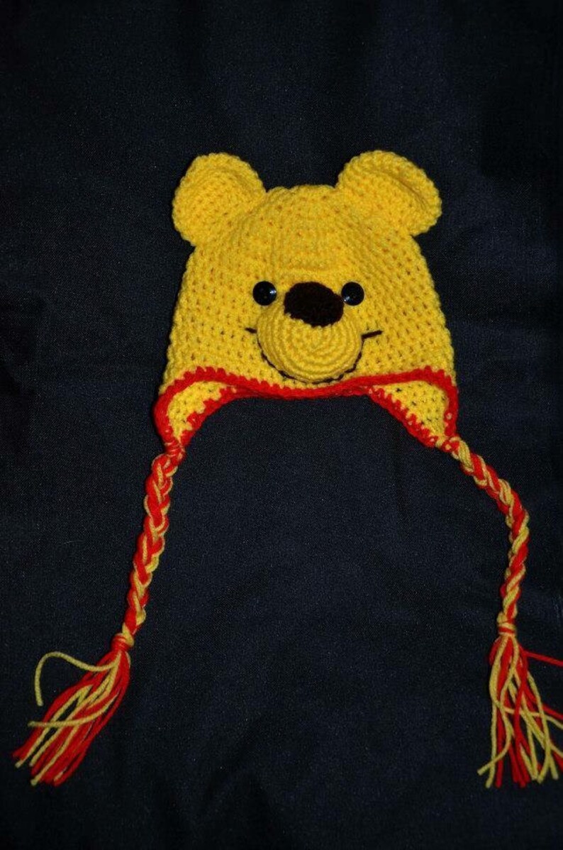Pode incluir: Um gorro de croch&ecirc; amarelo com design de rosto de urso, duas orelhas arredondadas, um nariz castanho escuro e olhos de bot&atilde;o pretos. O gorro inclui uma borda vermelha na parte inferior e duas longas tran&ccedil;as de l&atilde; amarela e vermelha.