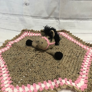 May include: A crochet security blanket featuring a tan horse head with a black mane, black hooves, and a pink halter. The blanket body is tan with a decorative border of alternating pink and white stitches in a classic granny square pattern.