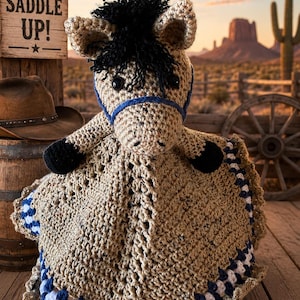 May include: A tan crochet horse lovey featuring a black yarn mane, black hooves, and a blue yarn bridle. The body consists of a textured, square-shaped blanket with a decorative border of white and navy blue yarn stitches.