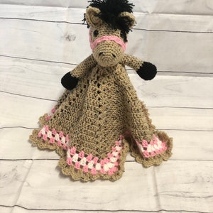 May include: A crochet security blanket featuring a tan horse head with a black mane, black hooves, and a pink halter. The blanket body is tan with a decorative border of alternating pink and white crochet stitches in a classic granny square pattern.