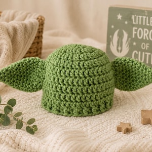 May include: A green crochet hat featuring two large, pointed ears extending from the sides. The hat has a textured, looped stitch pattern and is positioned near a wooden sign that reads "LITTLE FORCE OF CUTE" with a graphic of a creature.