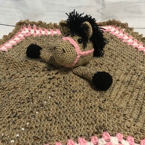May include: A crochet security blanket featuring a tan horse head with a black mane, black hooves, and a pink halter. The blanket body is textured tan yarn with a border of pink and white granny square stitches, creating a soft, square-shaped item.