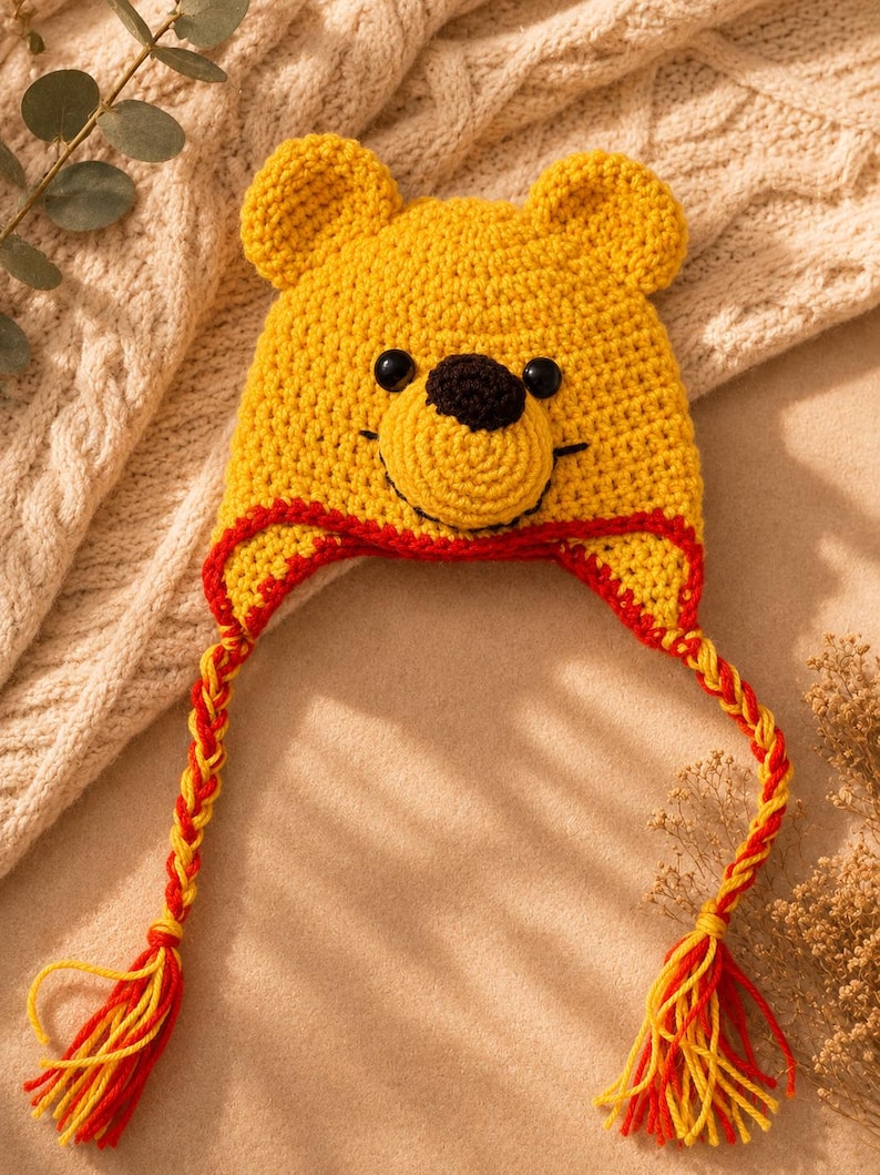 Pode incluir: Um gorro de croch&ecirc; amarelo com orelhas de urso, um nariz castanho escuro e olhos de bot&atilde;o pretos. O gorro tem uma borda vermelha e duas abas longas tran&ccedil;adas de fio vermelho e amarelo, cada uma terminando em uma borla de fio multicolorida.