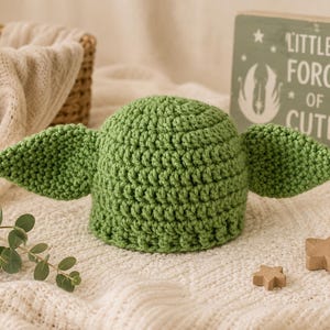 May include: A green crochet hat featuring two large, pointed ears extending from the sides. The hat has a textured, looped stitch pattern and is positioned near a wooden sign that reads "LITTLE FORCE OF CUTE" with a graphic of a creature.