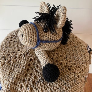 May include: A tan crochet horse lovey featuring a textured, speckled yarn body, a black mane, and black hooves. The horse head is adorned with a blue yarn halter and dark, round eyes, attached to a soft, crocheted blanket base with a decorative border.