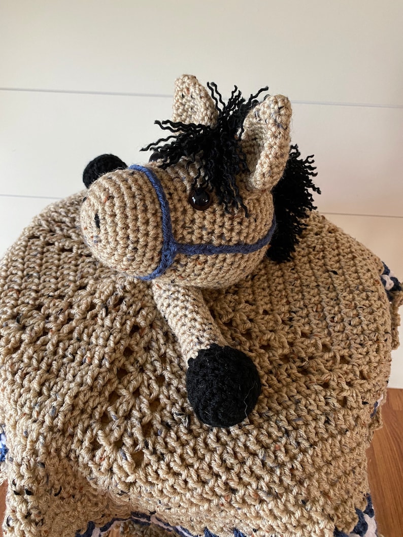 May include: A tan crochet horse lovey featuring a textured, speckled yarn body, a black mane, and black hooves. The horse head is adorned with a blue yarn halter and dark, round eyes, attached to a soft, crocheted blanket base with a decorative border.