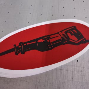 May include: An oval-shaped sticker featuring a bold red background with a black graphic illustration of a reciprocating saw centered horizontally. The sticker has a clean white border surrounding the red oval shape, creating a high-contrast design.