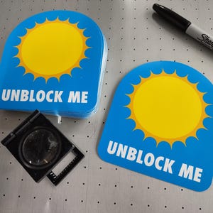 Puede incluir: Pegatinas azules con forma de arco, diseño de sol amarillo y las palabras "UNBLOCK ME" en blanco. También hay un rotulador Sharpie negro y una lupa.