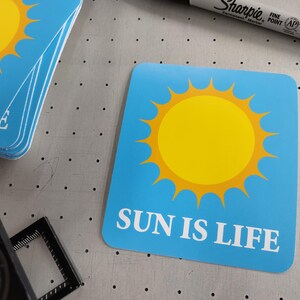 Puede incluir: Tarjetas azules con esquinas redondeadas, cada una con un gráfico de sol amarillo y el texto blanco "SUN IS LIFE". Un marcador negro es visible en el fondo.