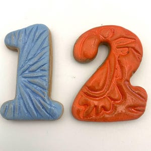 Może przedstawiać: Ceramiczna liczba "12" z niebieską i pomarańczową glazurą. Liczba "1" ma wzór gwiazdy, a liczba "2" ma wzór kwiatowy.